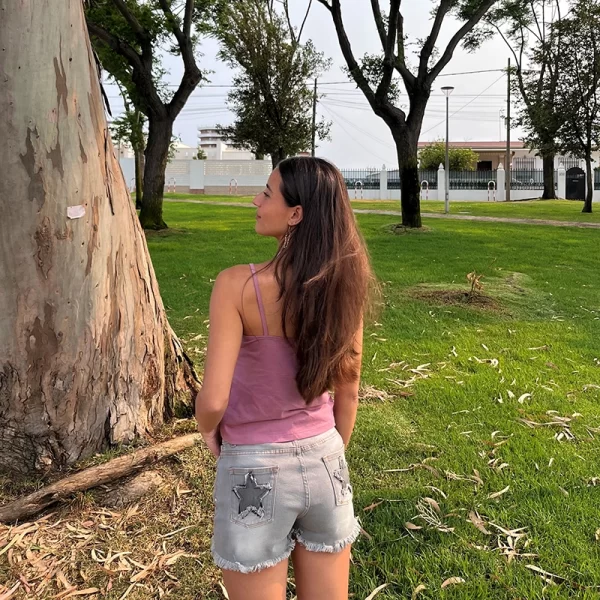 chica morena de espaldas posando en un jardín con un top de tirantes rosa y un short gris