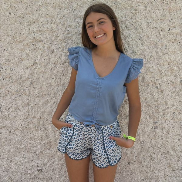 Irene Silva delante de una pared gris con un short estampado azul con fondo blanco con vivos de bolitas azules y una blusa azul con túnel frontal para fruncir cordón y tejido de agujeros en volantes de mangas cortas