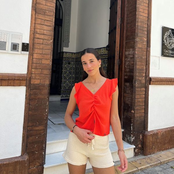 chica delante de una puerta abierta de un edificio con una blusa de manga corta en color coral y un short de tricotosa en crudo