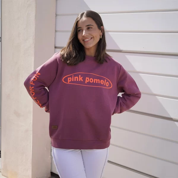 chica posando con una sudadera berenjena con estampado frontal con texto pink pomelo en naranja flúor y estampado juice for fun en manga derecha combinado con pantalón blanco