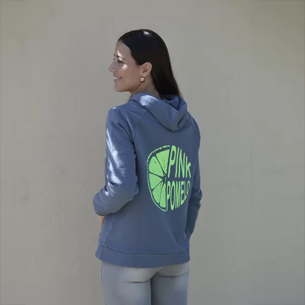 chica de espalda con Sudadera azul con capucha y bolsillo canguro frontal. Cordón azul personalizado Pink Pomelo en verde flúor en capucha, bordado verde flúor en bolsillo canguro frontal y print de rodaja de pomelo en pecho izquierdo y combinado con texto Pink Pomelo en espalda y un tejano azul claro