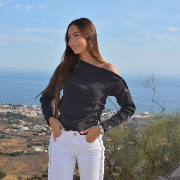 chica con un top antracita asimétrico con un hombro descolgado con cierres metálicos en lateral izquierdo y ambos brazos y pantalón blanco con flecos en laterales