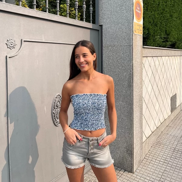 chica morena posando delante de una casa con un top banda estampado azul y blanco y un short gris