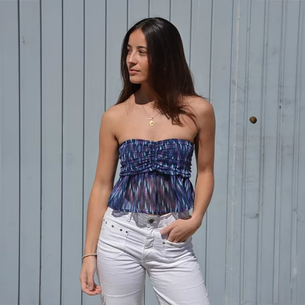 chica mirando hacia su lado derecho delante de un portón gris con un top palabra de honor en tonos azules y malvas con hilo de lúrex plateado y pantalón blanco con tachuelas