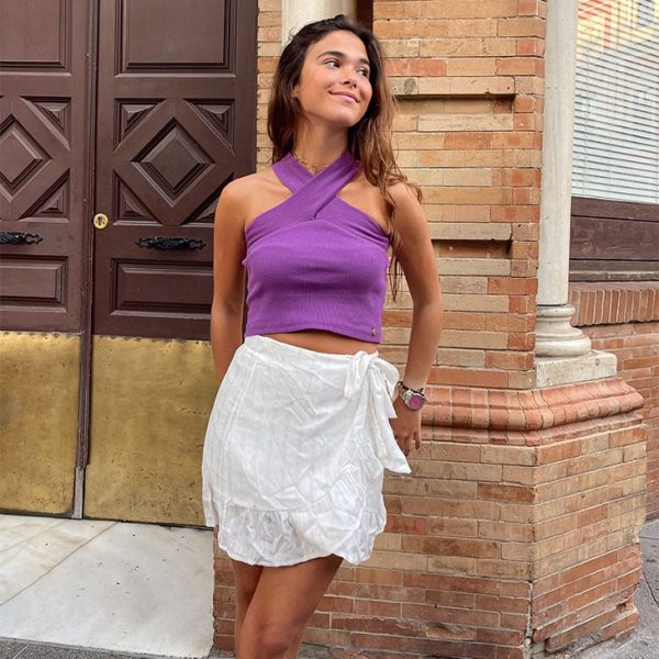 chica apoyada en la pared con un top morado corto cruzado al cuello y falda corta blanca