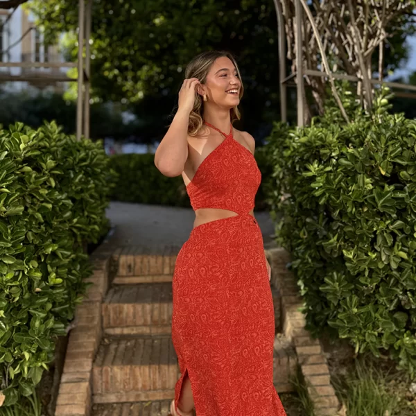 chica sonriendo bajando unas escaleras en un parque con un vestido largo cut out largo estampado en tonos coral y naranja con forro interior en coral. Hilos de lúrex a tono. Frunce nido de abeja en la espalda del top y de la cintura. Aberturas en ambos laterales del bajo.