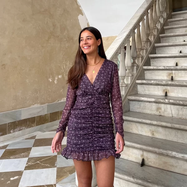 chica en un portal delante de unas escaleras con un vestido corto de gasa estampada en tonos negros, rosas, lilas e hilo de lúrex plateado de escote pico con manga larga, espada descubierta y forro interior