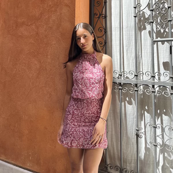 chica posando apoyada en una pared color albero con vestido corto cuello halter estampado en tonos rosas y blanco roto; con espalda descubierta. La parte de la falda es fruncida con goma de canilla. Forro interior de punto en color rosa